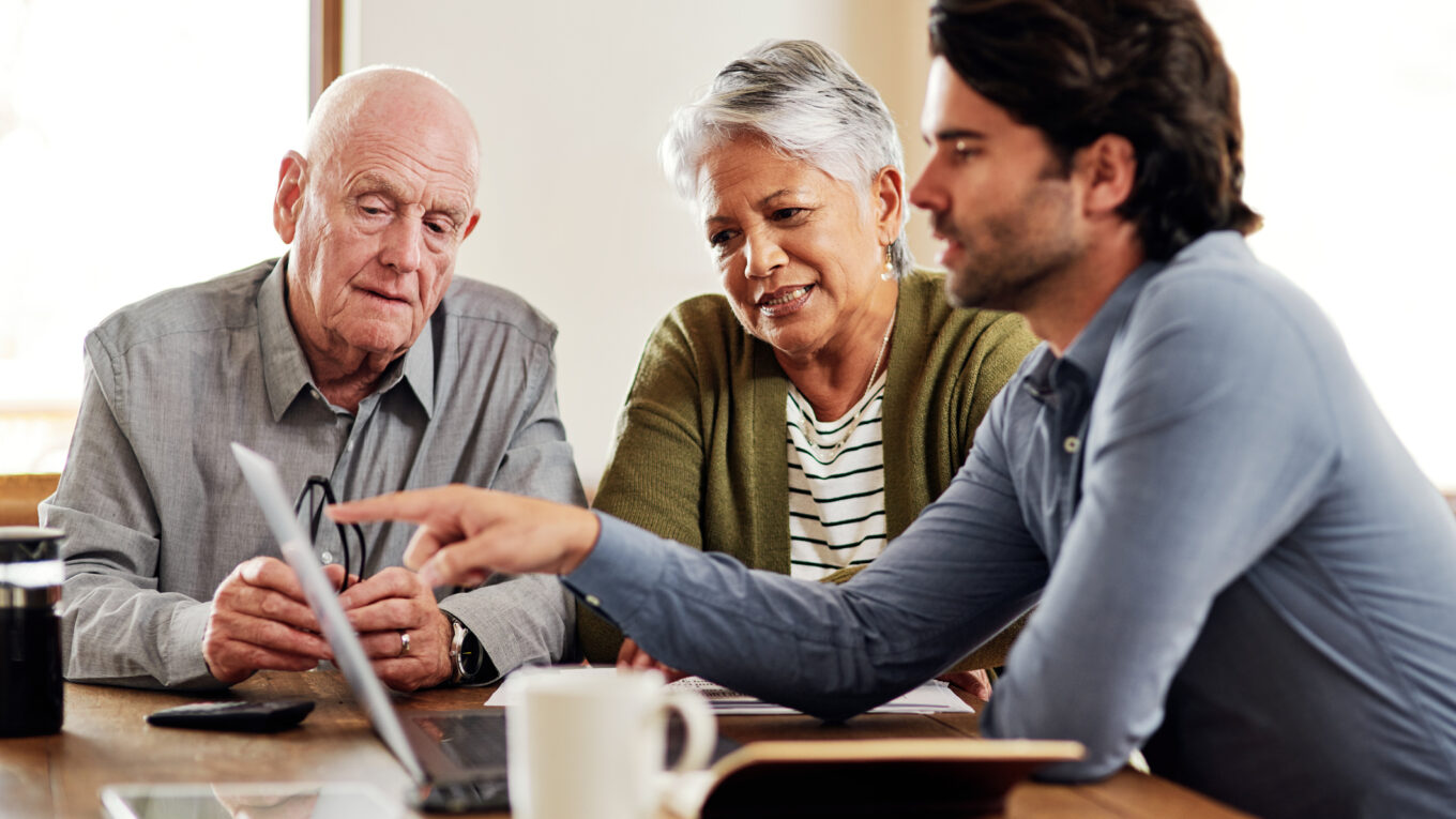 Two older people look at a laptop, and a younger man is pointing at the screen